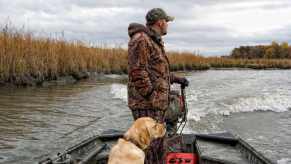 Man in camo clothing with a golden retriever at his feet pilots a small outboard boat.