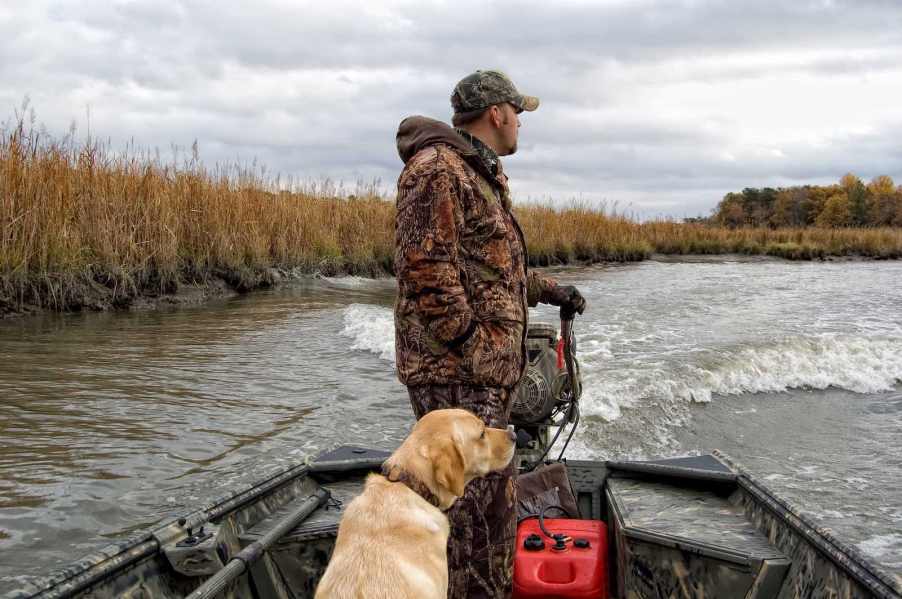 Man in camo clothing with a golden retriever at his feet pilots a small outboard boat.