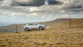 A light-colored Jaguar I-Pace electric SUV driving up a grassy hill remote location