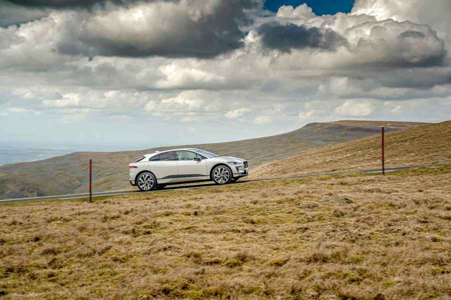 A light-colored Jaguar I-Pace electric SUV driving up a grassy hill remote location