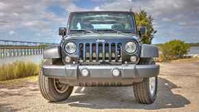 A black Jeep Wrangler parked near water with bridge in background in direct front view with front tires turned to left frame