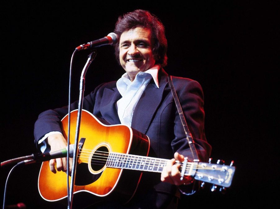 Johnny Cash smiles onstage holding guitar with black background in 1979 wearing white button up and dark jacket