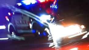 A blurred night scene of a police car with lights on and another car in front of it, illustration of how a Tesla driver using FSD mode ran into a police car in CA on June 14, 2024.