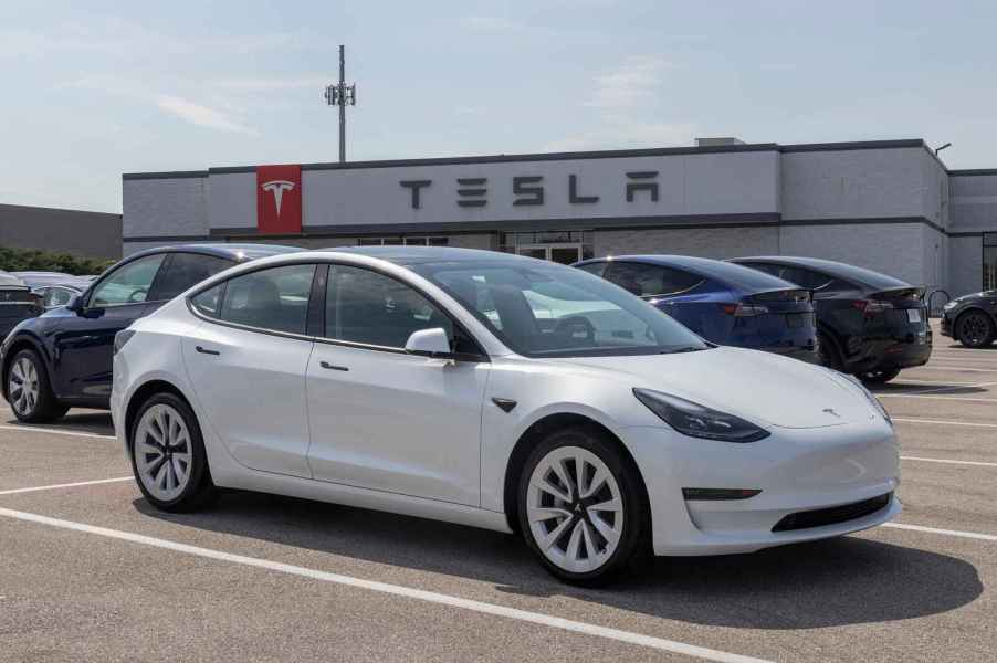 A white Tesla Model 3 EV sedan parked at a Tesla dealership parking lot spot in right front angle view