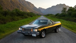 A dark-colored 1975 Ford Ranchero parked on mountainous gravel road in left front angle view with a thick yellow and orange stripe down the side