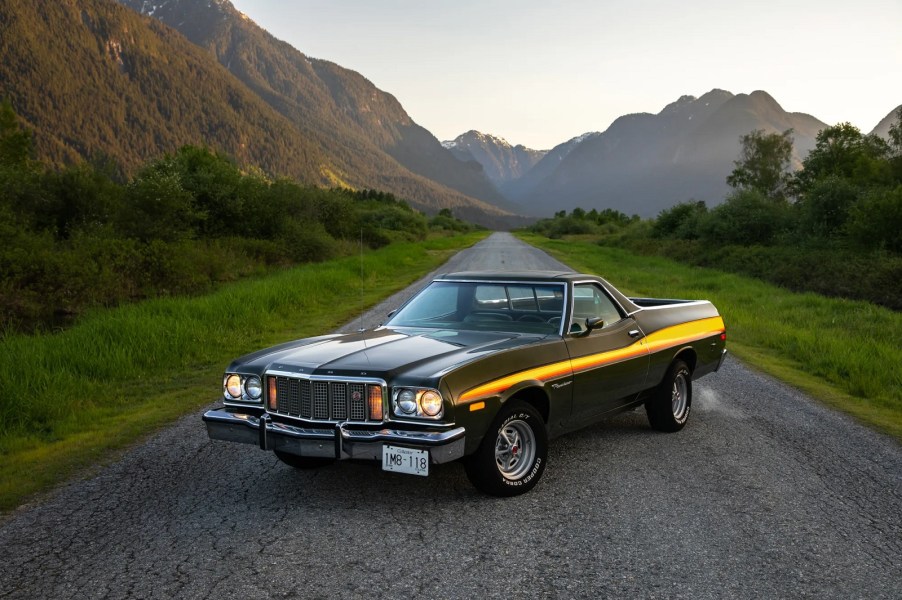 A dark-colored 1975 Ford Ranchero parked on mountainous gravel road in left front angle view with a thick yellow and orange stripe down the side