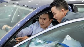 A customer sits behind the wheel of a new car while a salesman explains things.