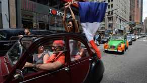 Young woman stands up in the back of a Citroën 2CV driving through Times Square, holding a French flag