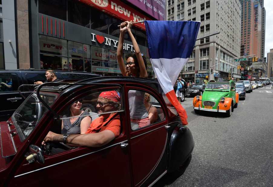 Young woman stands up in the back of a Citroën 2CV driving through Times Square, holding a French flag