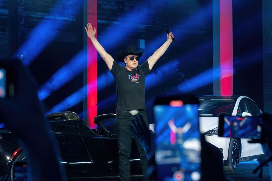 Elon Musk, wearing a belt buckle and cowboy hat, holds his hands aloft on a Tesla gigafactory stage while a lightshow goes on behind him.