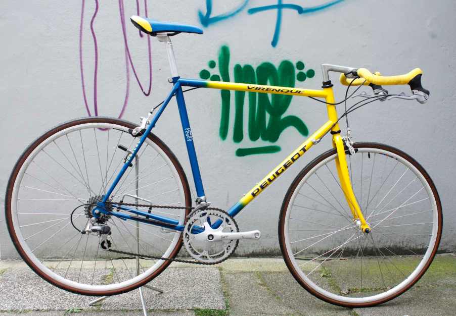 Vintage blue and yellow Peugeot road racing bicycle leaned against a graffiti-covered wall.