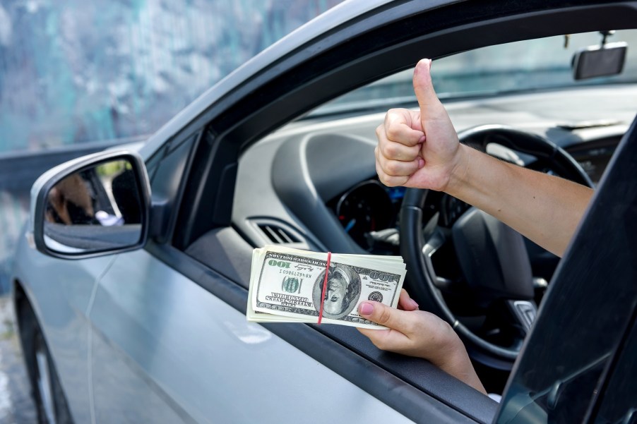 A depiction of a vehicle diminished value claim with a woman's hands holding a stack of money in left hand with right hand doing 'thumbs up' motion out of an open car window