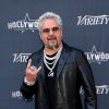 Guy Fieri stands on a red carpet doing a hand signal wearing multiple diamond necklaces and sunglasses