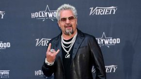 Guy Fieri stands on a red carpet doing a hand signal wearing multiple diamond necklaces and sunglasses