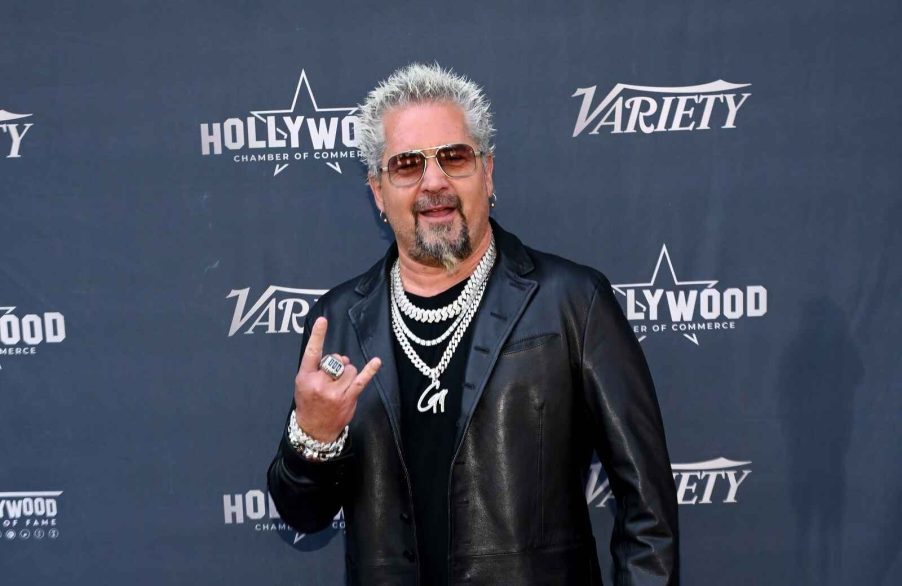 Guy Fieri stands on a red carpet doing a hand signal wearing multiple diamond necklaces and sunglasses