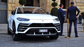 A white Lamborghini Urus SUV parked in slight right front angle view with two suited men standing facing away from the driver's side