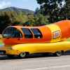 Oscar Mayer Wienermobile in left profile view on a flat paved road with grass and wooded hills in the background