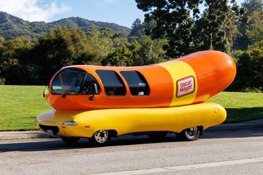 Oscar Mayer Wienermobile in left profile view on a flat paved road with grass and wooded hills in the background