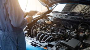 A mechanic performs a vehicle maintenance inspection on a blue car with hood open using an iPad