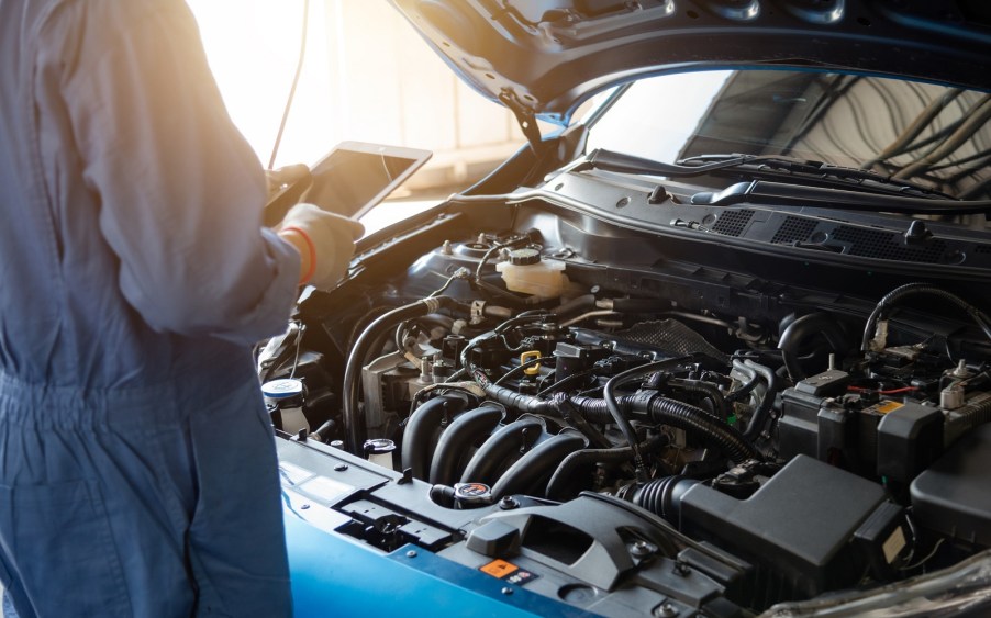 A mechanic performs a vehicle maintenance inspection on a blue car with hood open using an iPad