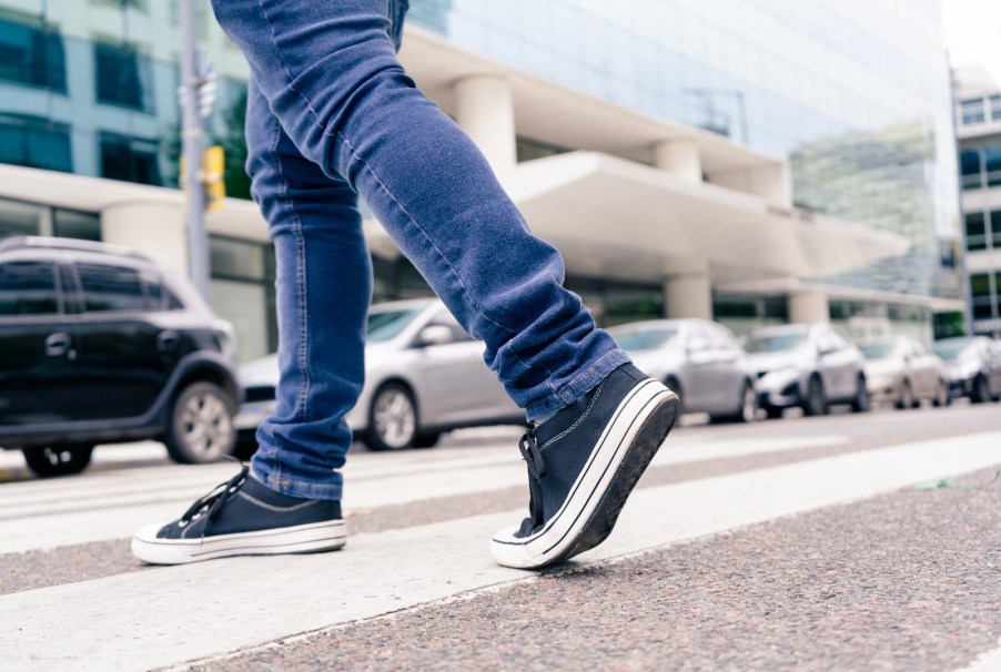 Depiction of stranger's legs wearing jeans and sneakers crossing the street approaching cars.