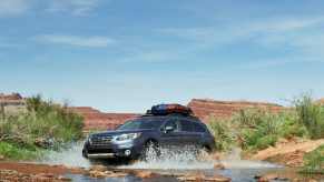 A Subaru Outback drives through a stream, mountains visible in the background.