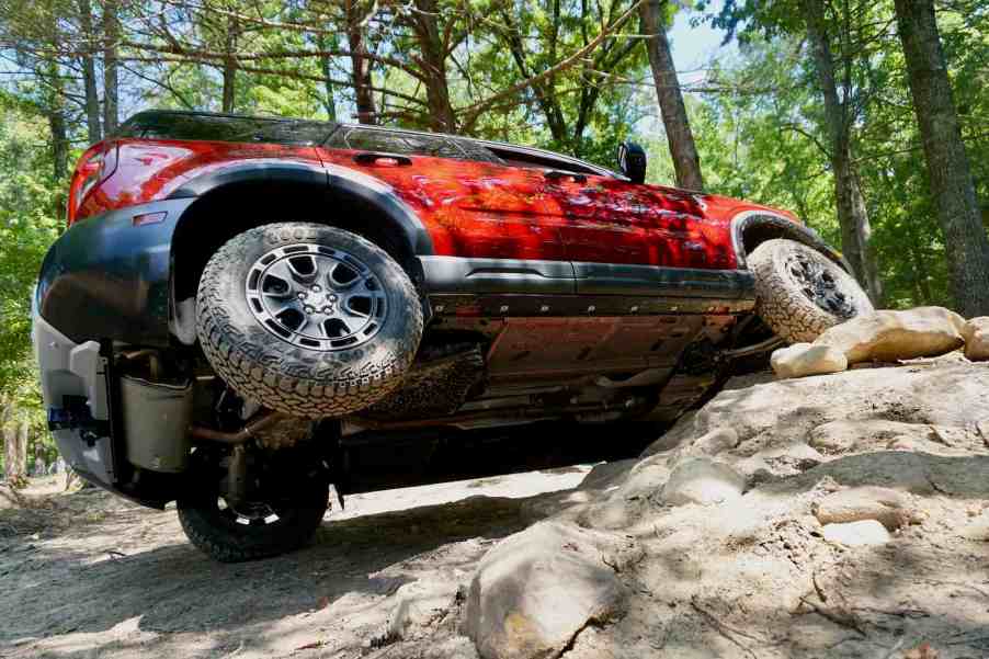 Red 2025 Bronco Sport Badlands shows off its 29-inch Goodyear tires by lifting one in the air as it crawls over rocks, trees visible in the background.