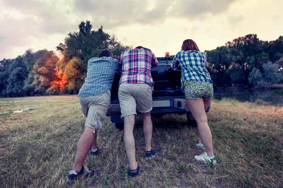 Group of guys push a broken down pickup ruck through a field, trees visible in the background.