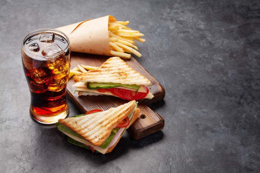 Club sandwich and a cold, iced soda sitting on a table, french fries visible in the background.