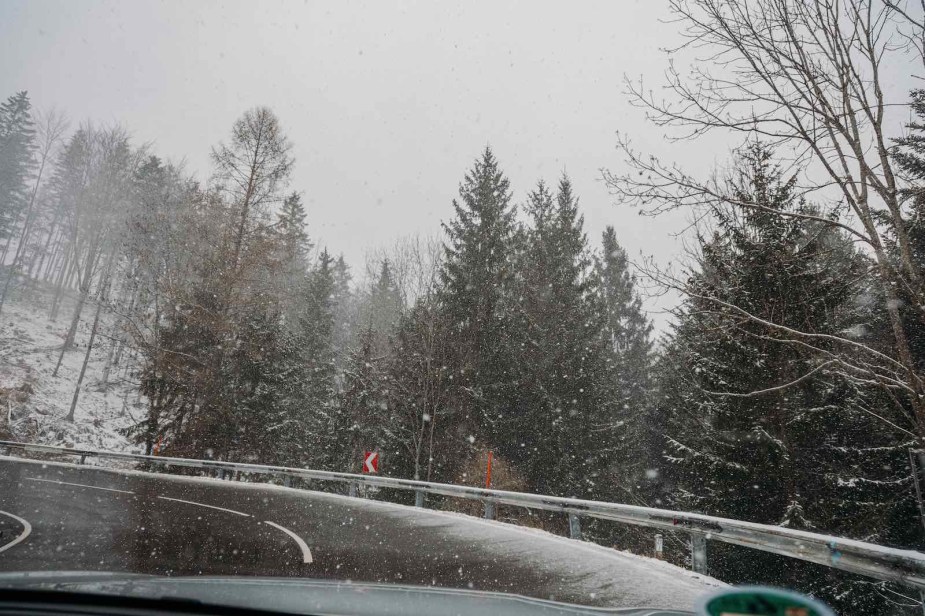 Snowy road winding up a mountain, trees visible in the background.