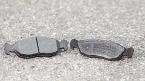 Two car brake pads, one new and one old, placed side by side on pavement in close view