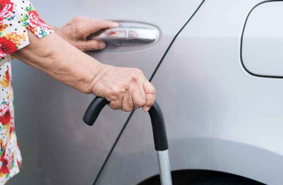 An elderly person's hand on a car door handle and cane in the other