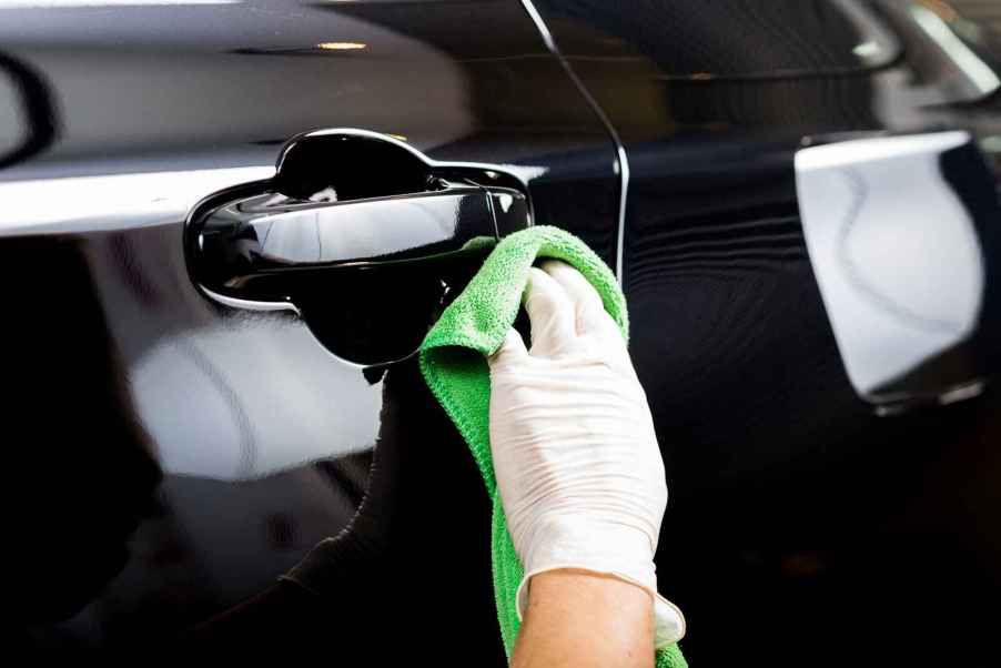 Car detailer hand wearing white latex glove wiping black car exterior door with a green microfiber cloth in close view