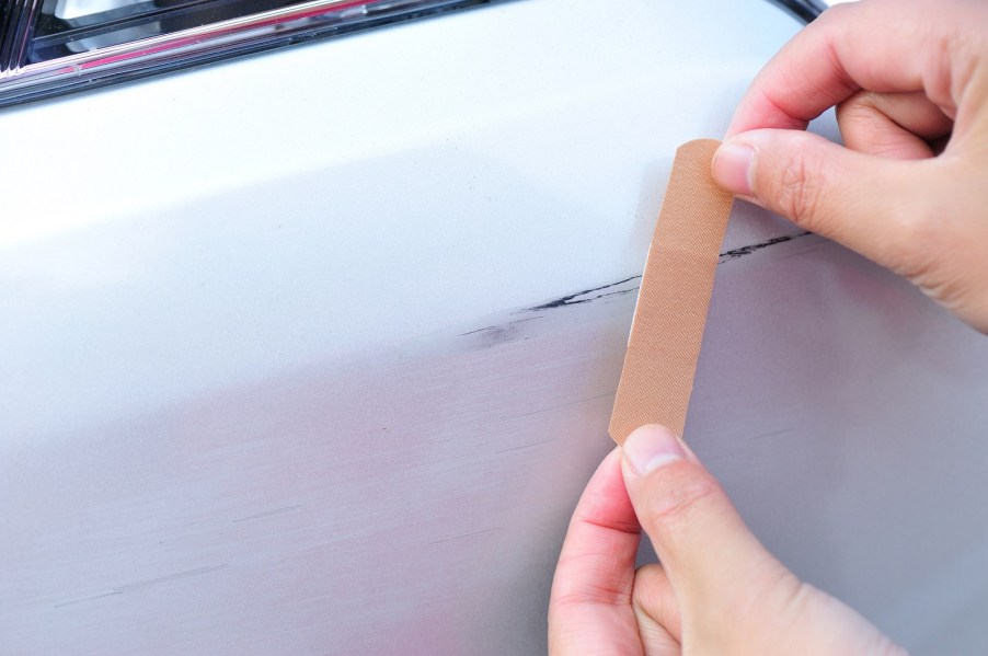 A scratch on a car in close view with hands placing a band-aid over it you might consider avoiding a car insurance claim for small damage