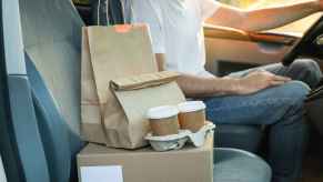 A driver for a food delivery service such as Uber Eats shown neck-down sitting in the car with packaged food orders on the passenger seat