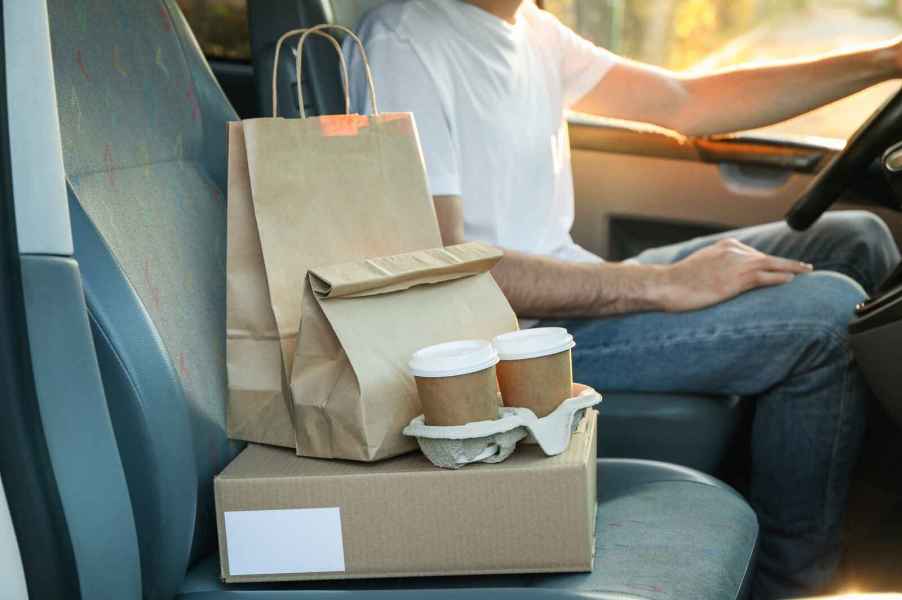 A driver for a food delivery service such as Uber Eats shown neck-down sitting in the car with packaged food orders on the passenger seat