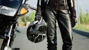 Motorcycle rider wearing leather gear holding a helmet shown from torso to shins