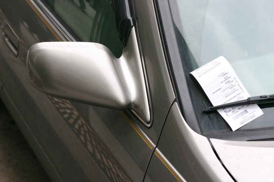 A parking ticket on a silver car's windshield tucked under the right wiper blade in close view