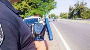 Police offer stands in front of police car parked on side of road holding radar gun