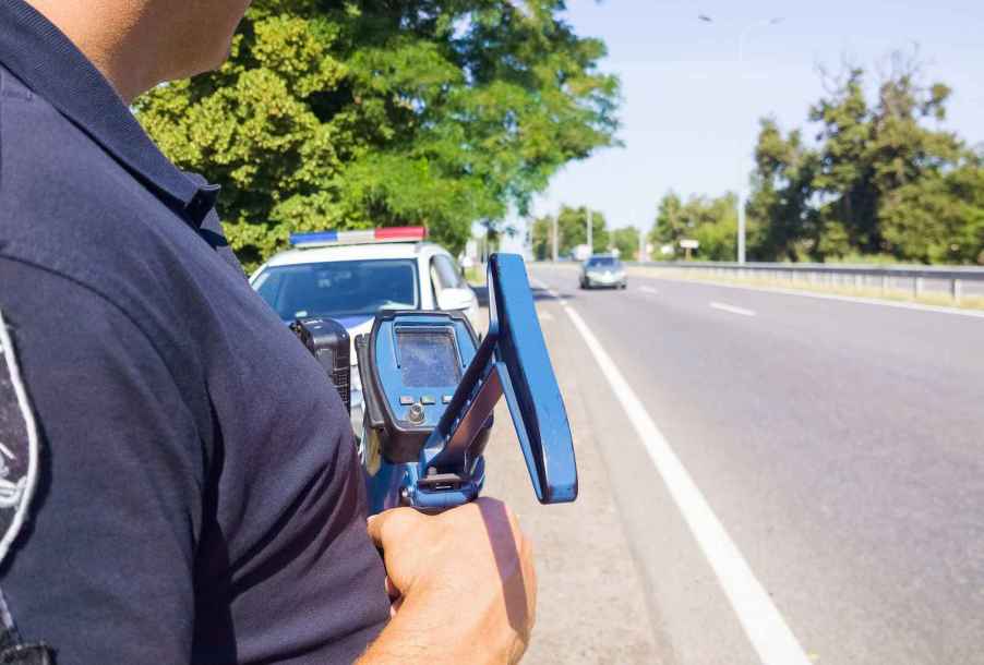 Police offer stands in front of police car parked on side of road holding radar gun