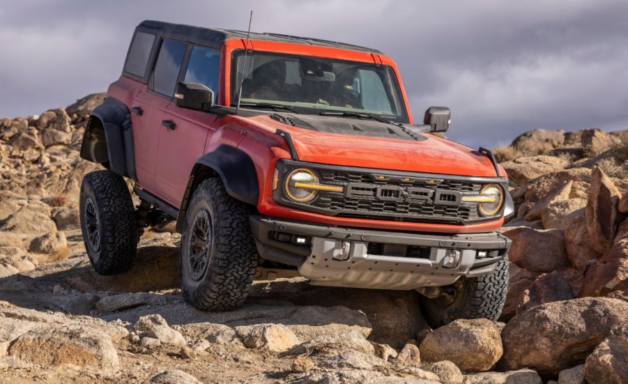 A reddish-orange 2022 Ford Bronco Raptor SUV climbing dusty boulders off-road in right front angle view