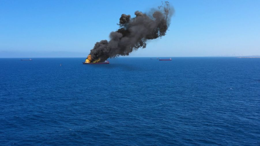 Column of black smoke coming from a burning yacht on the ocean, a blue sky visible in the background.