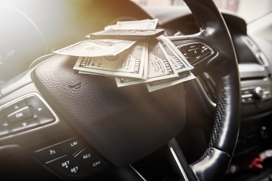 Stack of $100 bills set on the steering wheel of a car.