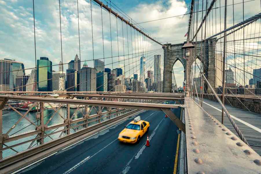 Bright yellow New York City taxi cab crosses the Brooklyn bridge, Manhattan's skyline visible in the background.