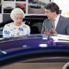 The Queen of England looks at a Jaguar car in surprise while the man next to her talks.