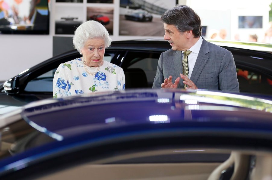 The Queen of England looks at a Jaguar car in surprise while the man next to her talks.