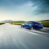 A blue Tesla Model 3 kicks up water on the road surface.