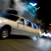 White limousine driving through a city at night, a blur of lights visible in the background.