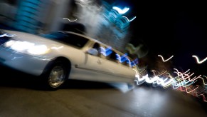 White limousine driving through a city at night, a blur of lights visible in the background.