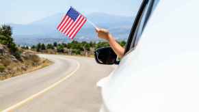 Hand holding small American flag out of white car driving on the highway in close side view